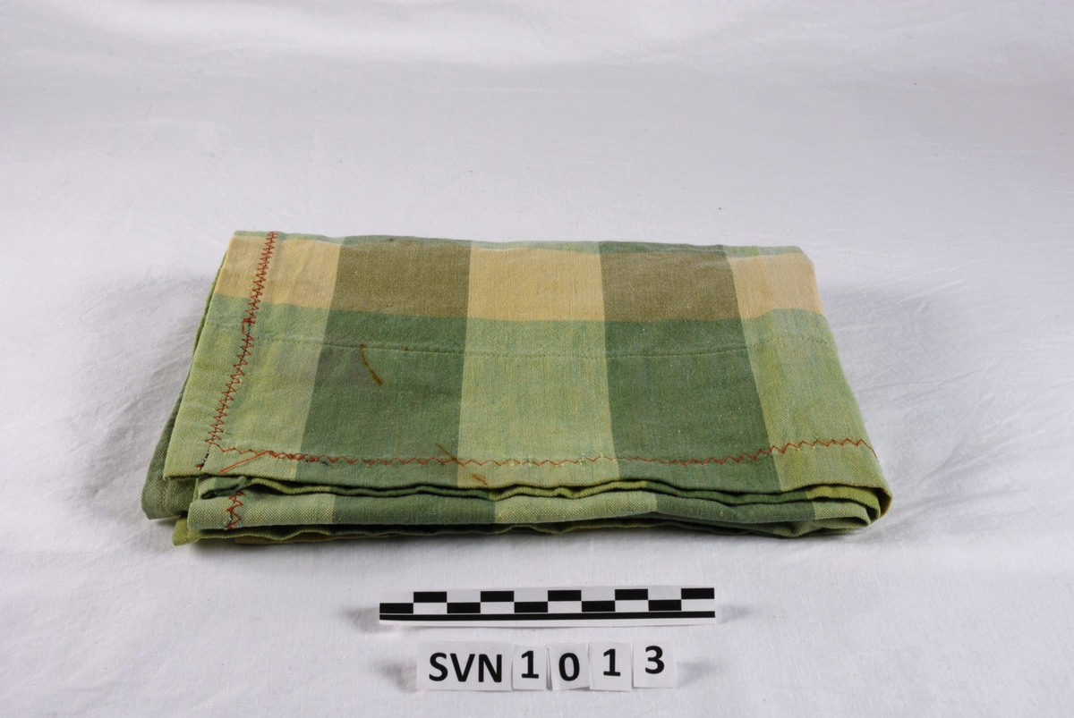 Grønn og gulrutete tekstilstykke. Alle sidene er foldet inn og sydd. I tillegg er det sydd en sikksakksøm rundt hele duken et stykke innenfor kantene.