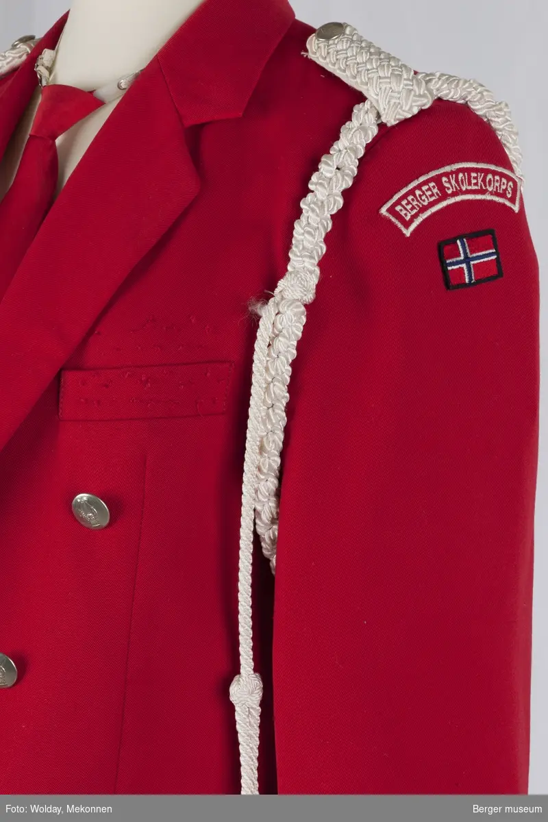 Åpen kort uniformsjakke i  rød stoff med silkefor i rødt, med hvit pyntesnor på den ene skulderen. Det er hvite epåletter i flettet silke.  Det er også et norsk flagg sydd på ventre arm under merket BERGER SKOLEKORPS. Knappene er dekorert med harper.