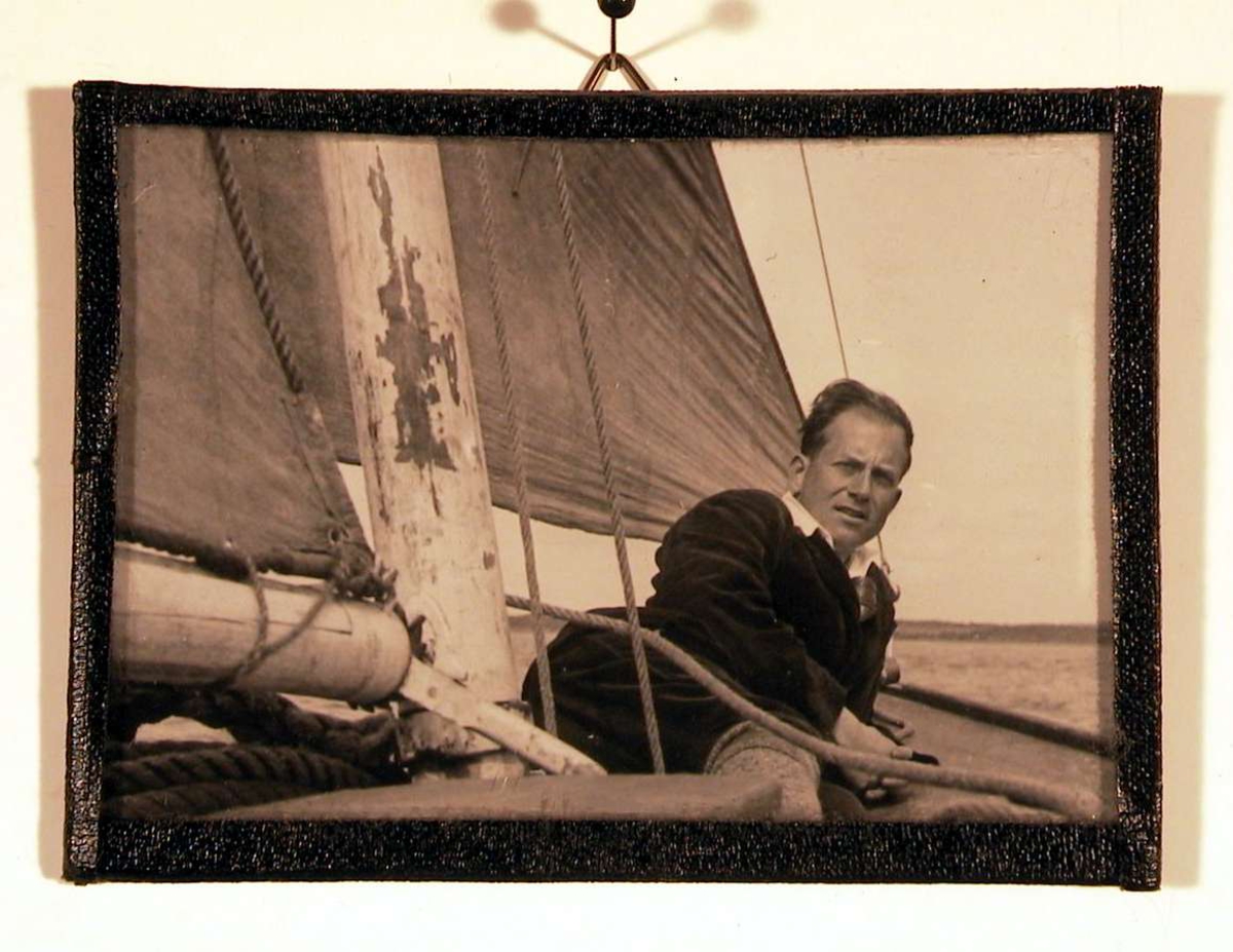 Fotografi av voksen ung mann liggende på dekket av gammeldags trebåt med kulørt seil. Han ser direkte på fotografen.
