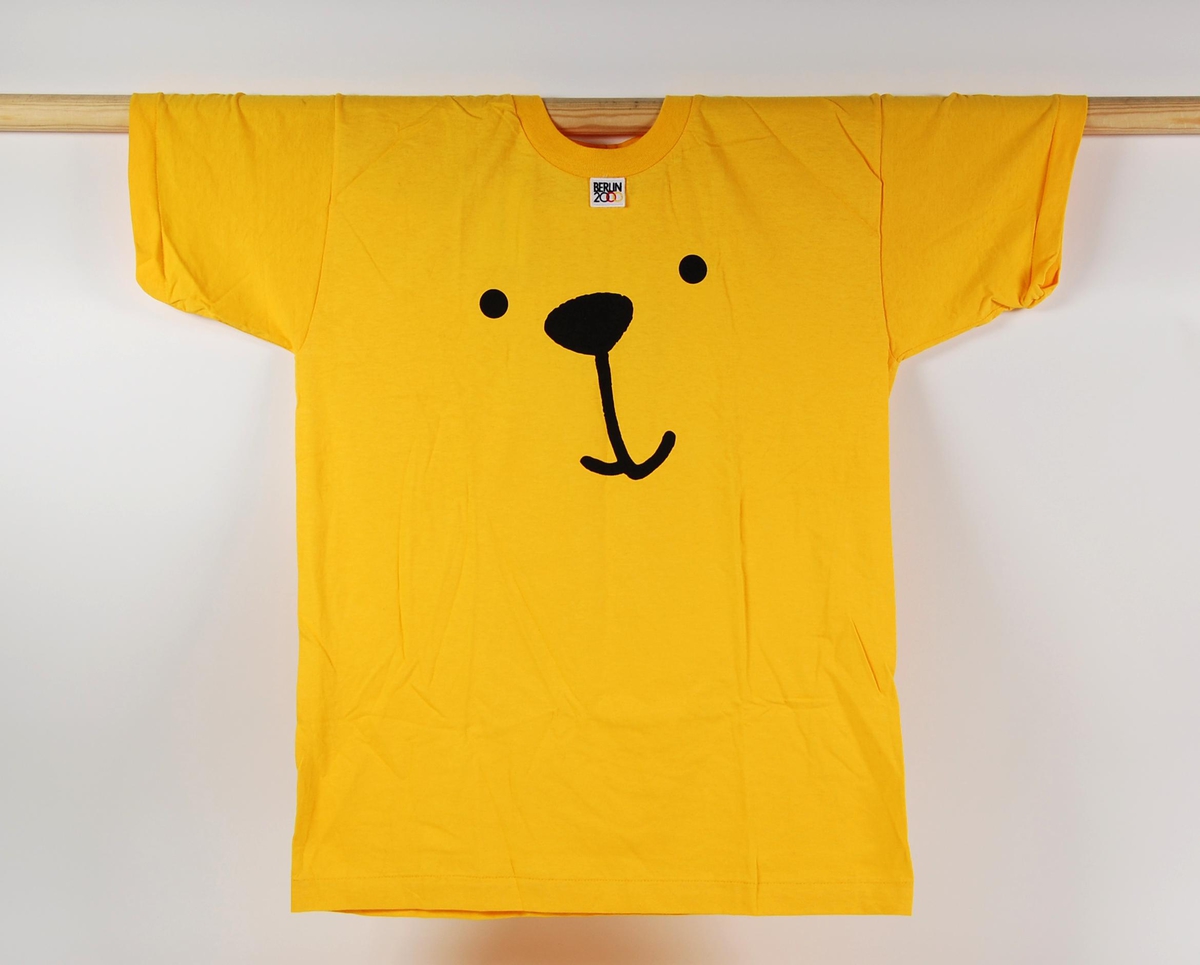 Gul og sort t-skjorte. Motivet på t-skjorten er ansiktet til en bjørn. Det er også påsydd et merke med innskriften: BERLIN 2000.