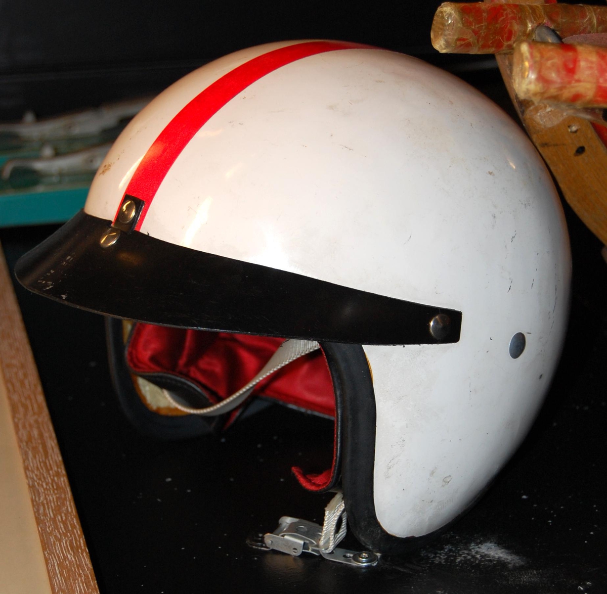 Hjelm av typen halvhjelm med kort skygge/skjerm. Hjelmen er hvit med en rød stripe. Skjermen/skyggen og kanten er sort