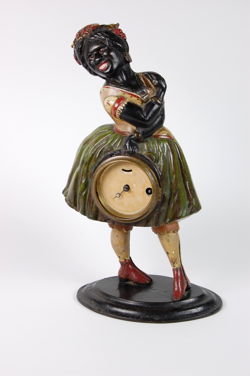 Støpt og malt figur formet som en afrikansk jente som bærer en klokke. Figuren har oval sokkel. Klokken er satt inn foran på skjørtet.