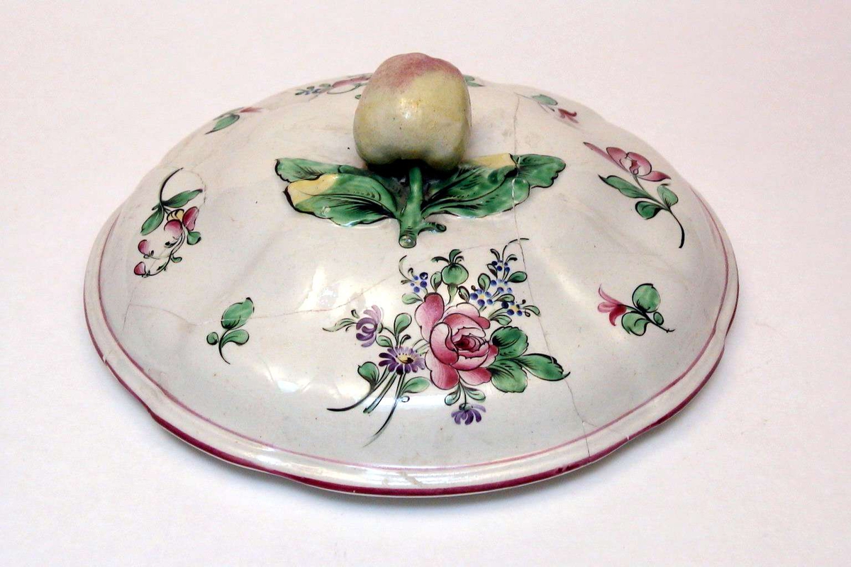Rundt lokk i kremfarget keramikk med polykrom blomsterdekor. Lokkgrepet er et eple. Lokket er defekt.
Fatet eller terrinen som lokket har hørt til, mangler.