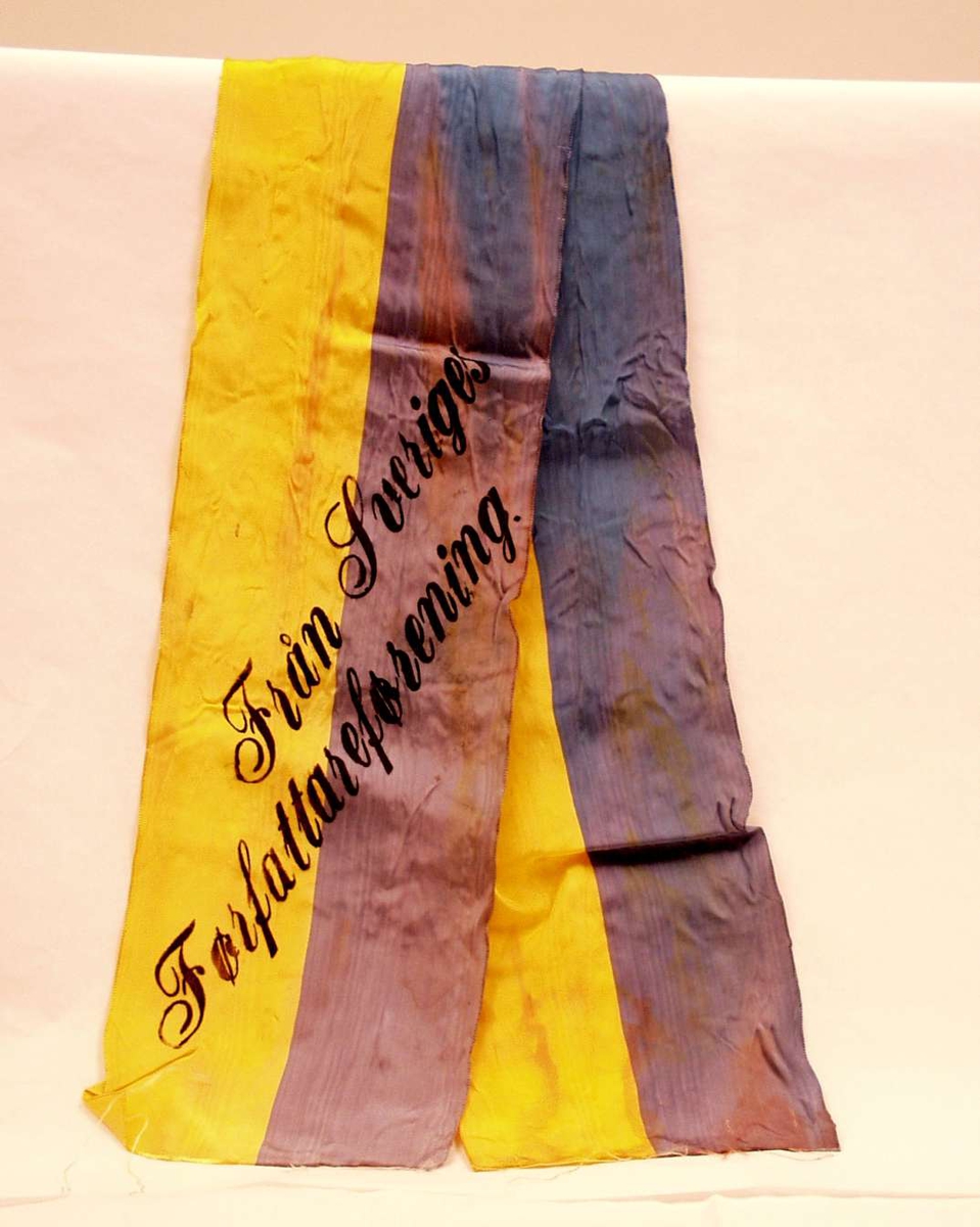 Bånd i gul og blå silke. Båndet er delt i to. På båndet står det: Från Sveriges Førfattareførening.