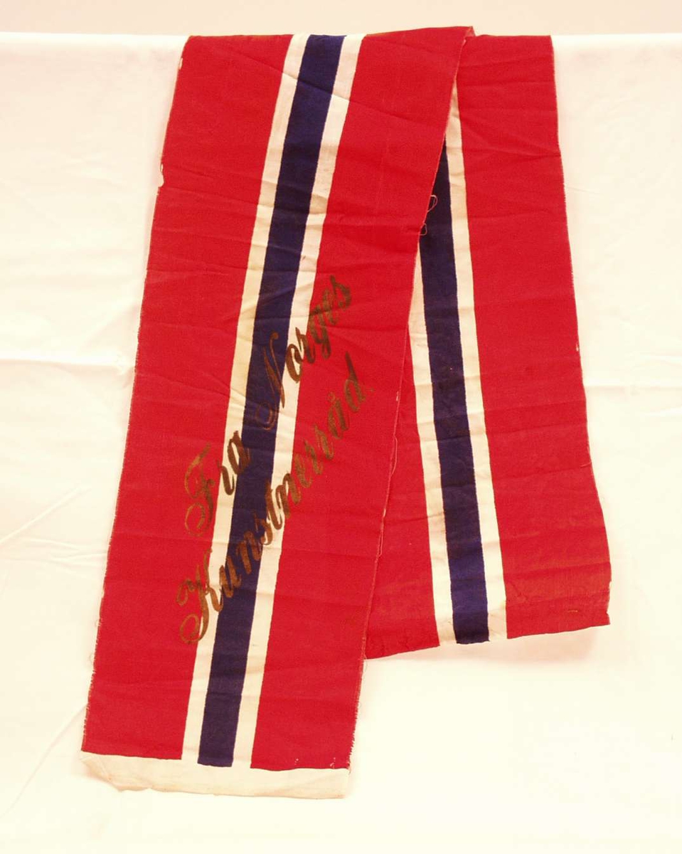 Bånd i rød, hvit og blå silke; flaggremse. Båndet er i to deler. På det ene står det i forgylte bokstaver: Fra Norges Kunstnerråd.