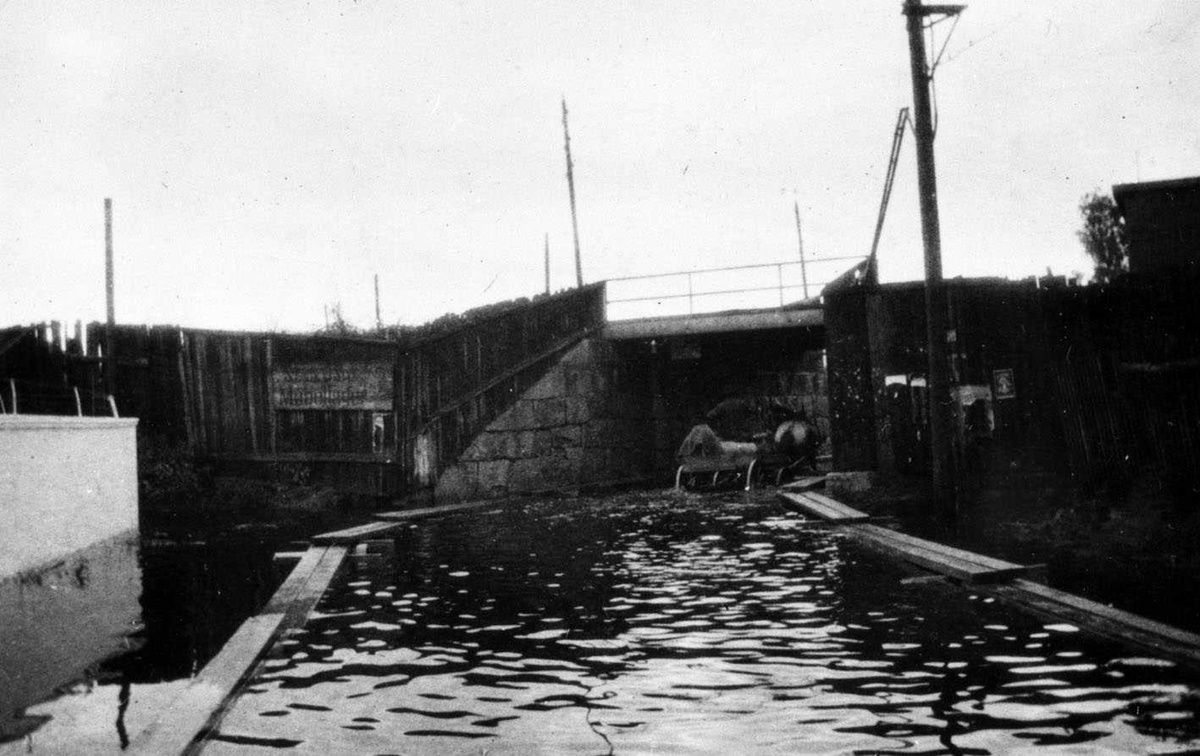 Jernbaneundergang i Lillestrøm under (regnværs?)flom 1910.
På begge sider av veien ligger flåteganger. En hest med trille vader gjennom undergangen. Vannet når omtrent til akslingen på trilla.