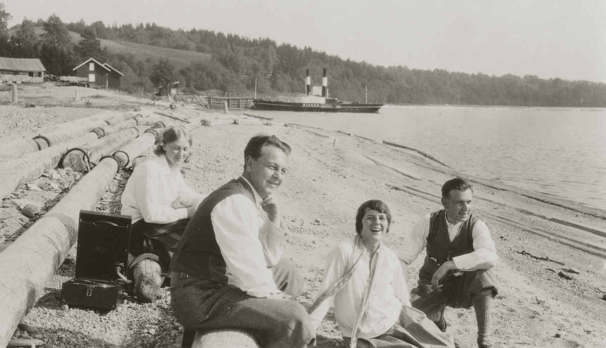 To kvinner og to menn sitter ved starndkanten. Fløte-tømmer til venstre. Portabel grammofon.