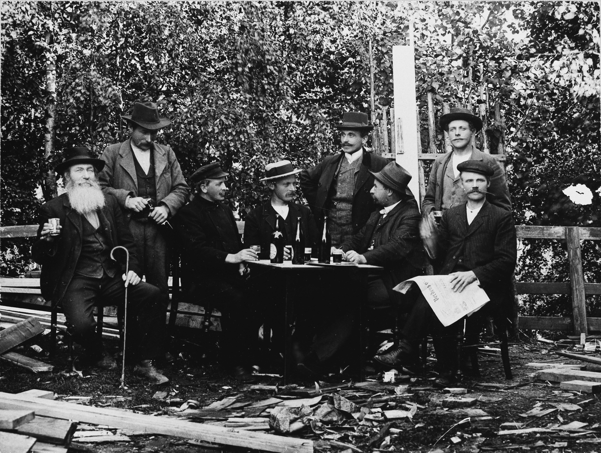 Gruppe med menn rundt et bord med hatter, ølflasker og avis.