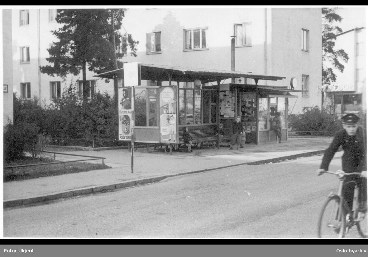 Aviskiosk og leskur / bussholdeplass med reklameplakater. Mann på sykkel i gata. Frisør-lokale i bakgrunnen til høyre. Stockholm, Sverige?