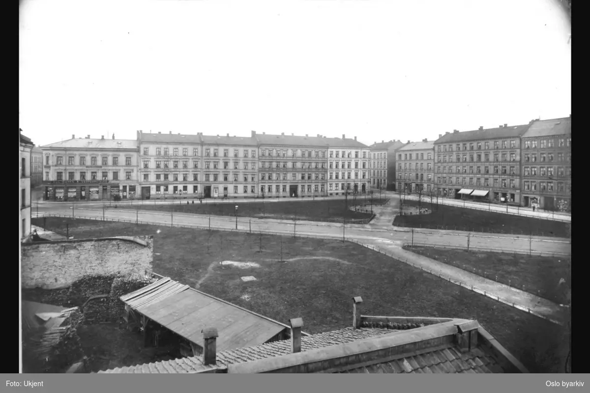 Parkområde på Schous plass med gangveier og plener. (Parklagt 1915 - 1916). Bygårder i Nordre gate (til venstre) og Toftes gate. Diverse butikker med reklame / skilt. Vedlager nærmest i forgrunnen. (Jfr. bilde A-20031/U/0014/070)