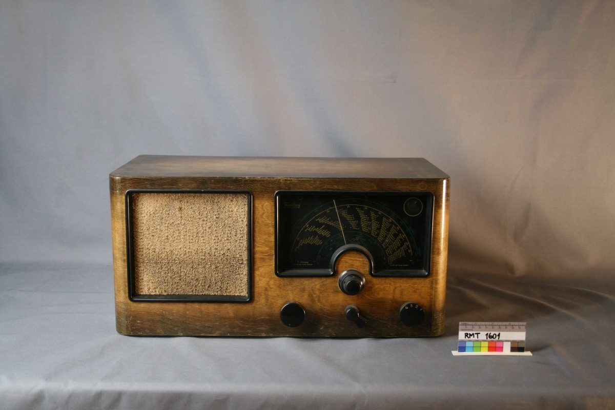 Rektangulær brun radio. Forside: høyttaler med tekstiltrekk, skala og fire funksjons-, og søkeknapper. Bakside: bakplate og strømuttak.