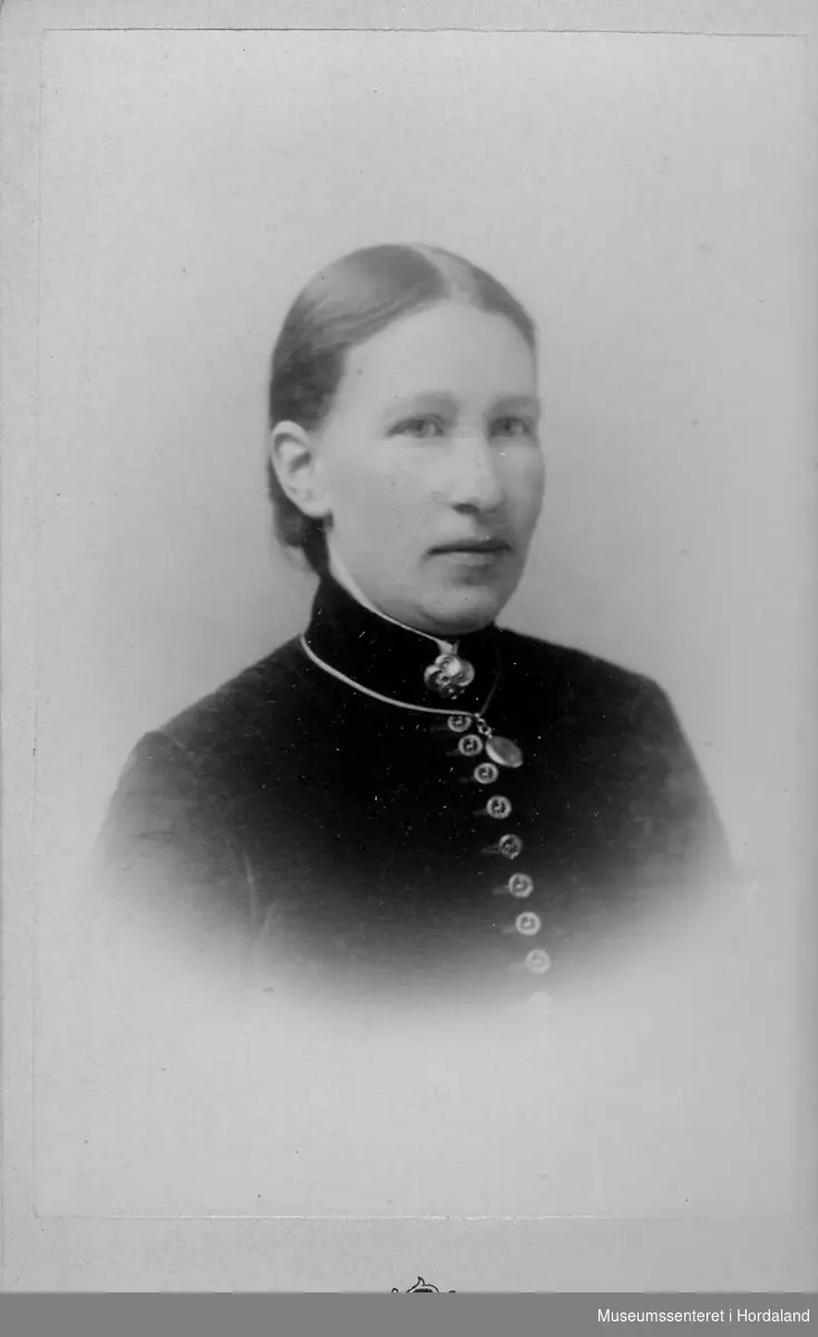 Ung kvinne med mørk jakke med høg hals, knappa framme med mange små knapper, sølje i halsen, medaljong rundt halsen.
