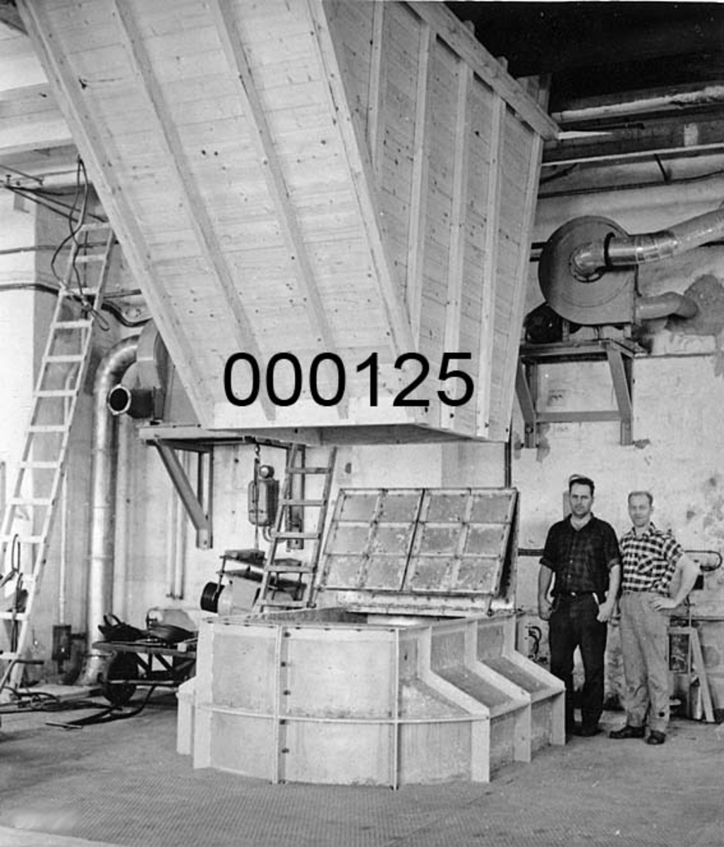 Interiørbilde fra Drammenselvens Papirfabrikker på Geithus på Modum i Buskerud. To karer ved den nye utskuddspulperen som sto ferdig i mars 1968.  Pulperen hadde typebetegnelsen "Jones Hi-Lo Pulper No 2 1/2".  Den hadde 11 kubikkmeters tank.  Tanken var at pulperen i hovedsak skulle løse opp utskuddspapir fra PM III, men rørsystemet var opplagt slik at den i nødsfall også kunne ta utskuddspapir fra PM IV.  Den erstattet en hollender som etter tradisjonen ved bedriften skulle ha vært innkjøpt til Skotfos Bruk i 1896 og overflyttet til Drammenselven i 1935. Informant Øyvind Haugen har ikke greid å identifisere mennene.  Pulperen står på golvet, mens ei sjakt av umalt trevirke henger ned fra taket over maskinen. 