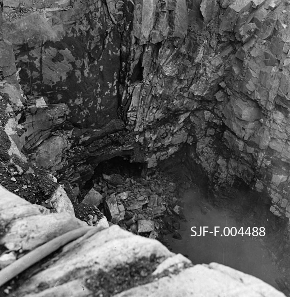 Gjennomslag for driftstunnelen til det som skulle bli Geithusfoss kraftverk på Modum i Buskerud.  Fotografiet er tatt 16. august 1960, fra ei berghylle ned mot ei sjakt hvor det står en del skittenbrunt vann, men hvor vi også aner en mørk åpning i fjellet.  Driftstunnelen var bortimot 110 meter lang, og gikk fra et inntakspunkt på østre elvebredd ovenfor fossen, under industrianlegget til Drammenselvens Papirfabrikker og til ei kraftverkstomt nedenfor fabrikken.  Dette fotografiet er antakelig tatt på den søndre sida.  Det tok et drøyt år fra tunnelgjennomslaget til kraftstasjonen ble satt i drift, 1. oktober 1961. 