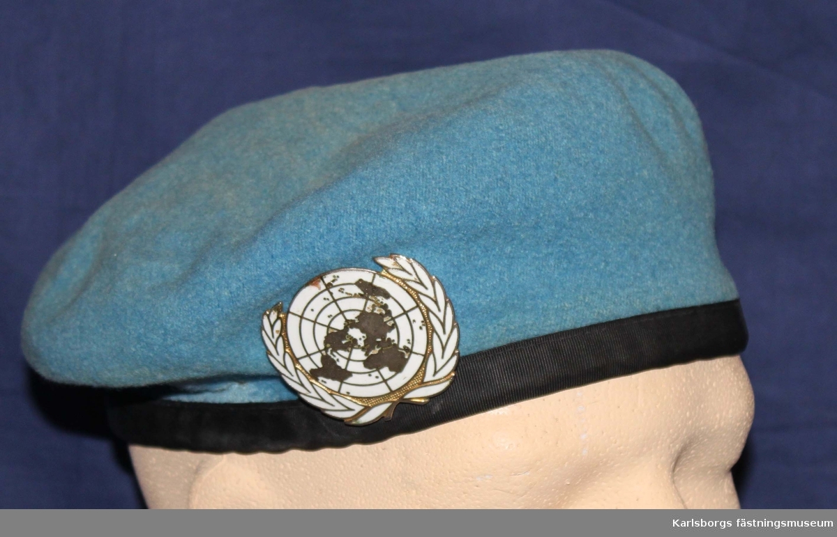 Mössa i baskermodell av ljusblå filt med skyddskant av skinn med inliggande dragband och försedd med baskertecken UN.