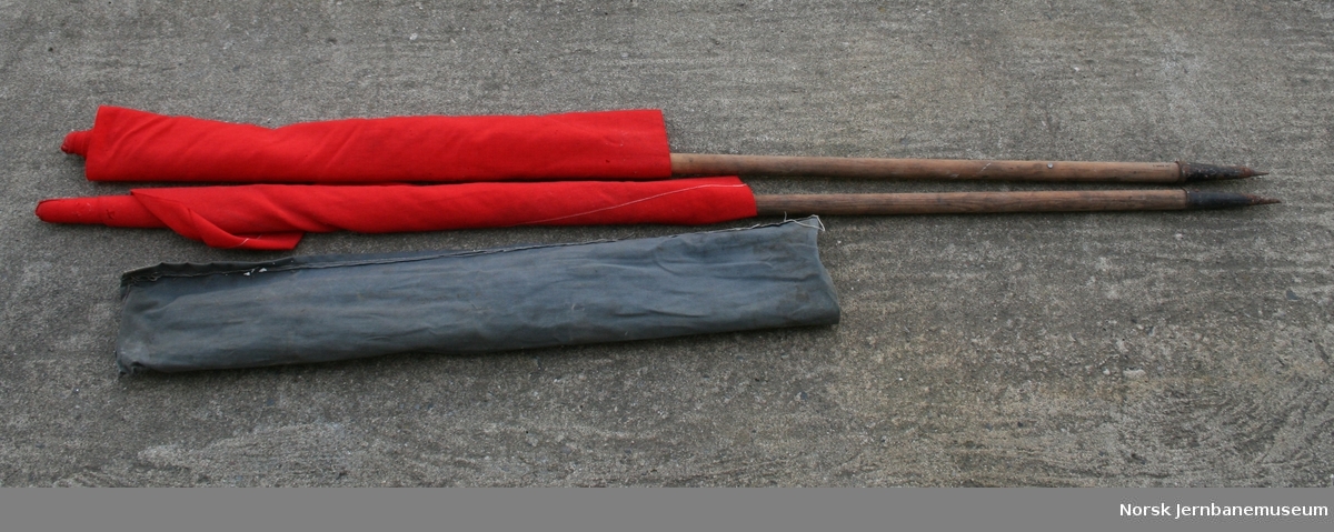 2 røde signalflagg i lerretspose. Festet på trestenger med metallspiss i enden. Brukt ved banearbeid.