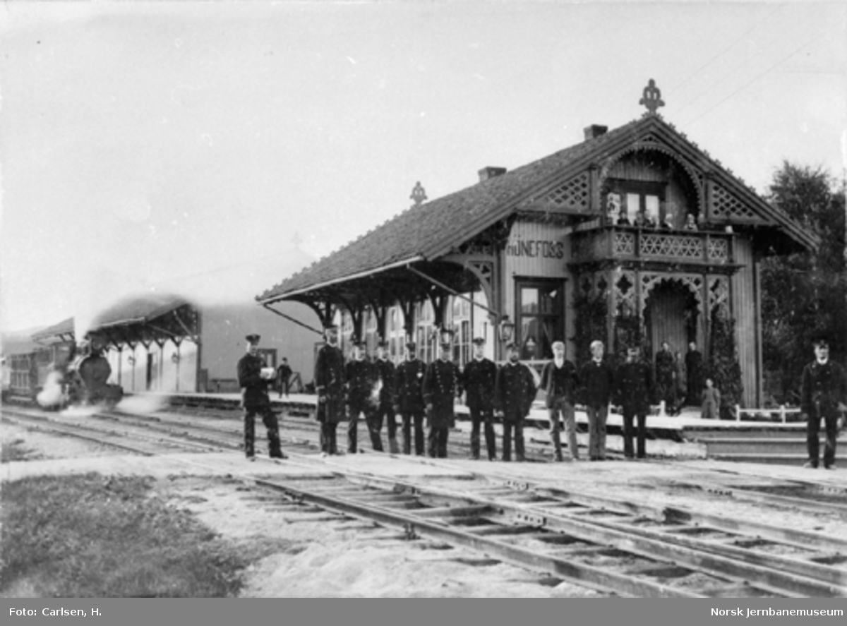 Personalet på Hønefoss stasjon oppstilt for fotografering med stasjonsbygningen og tog 184 "Slaven" i bakgrunnen