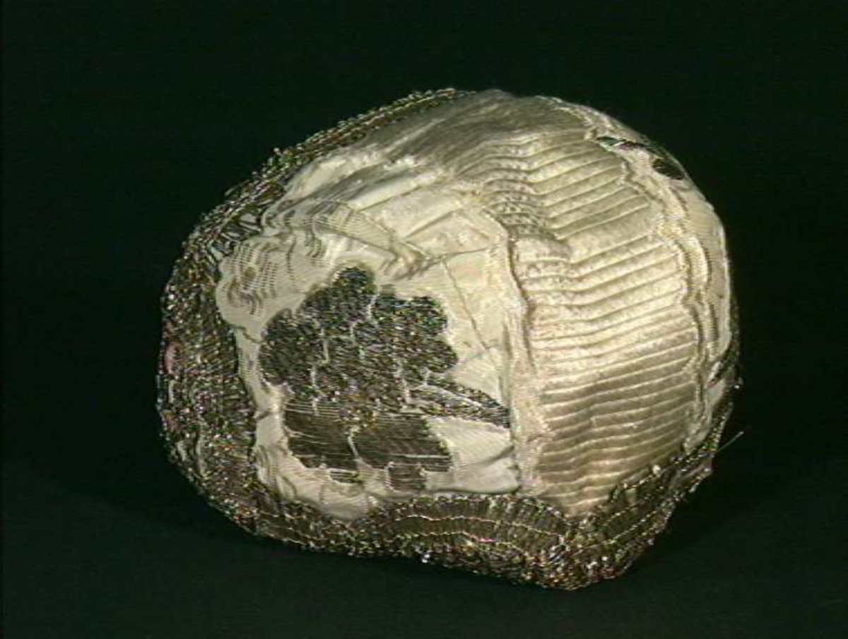 Hvit dåpslue i silke med brosjert mønster i metall og dekor av kniplet metallblonde. Rosa silkes fôr.