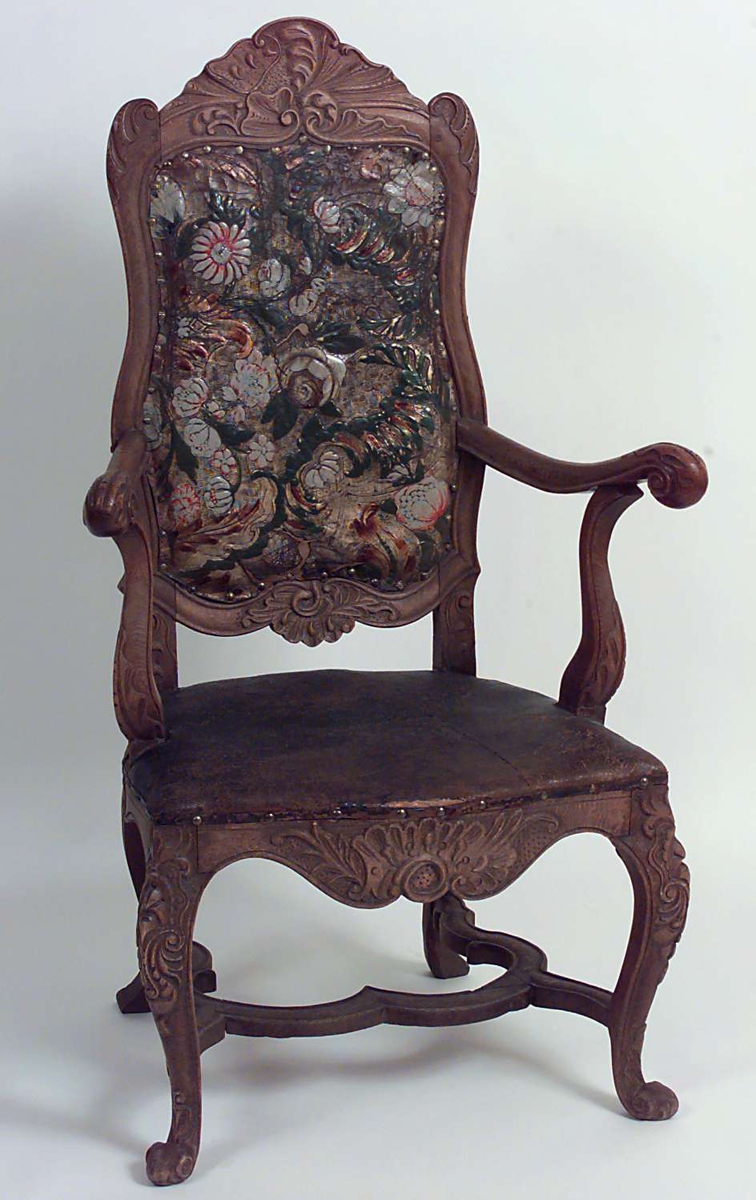 Regence-stol med ornamentikk i tre- og gyldenlærsarbeide som knytter den til rokokko-perioden. Gyldenlær trekk på stolryggen, blomsterdekor. Lær trekk på setet, brunt ensfarget. Armlener. S-formet ben. Utskåret dekor på armlener, ben, sarg, rundt setet og stolryggen.