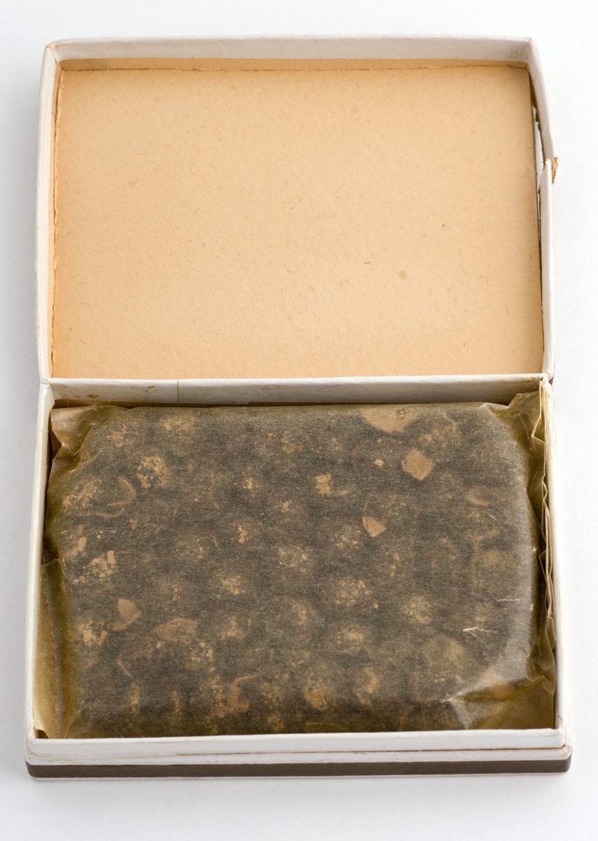 Firkantet eske av papp som inneholder brune tabletter støpt i en blokk. Tablettene er innpakket i gjennomsiktig matpapir.