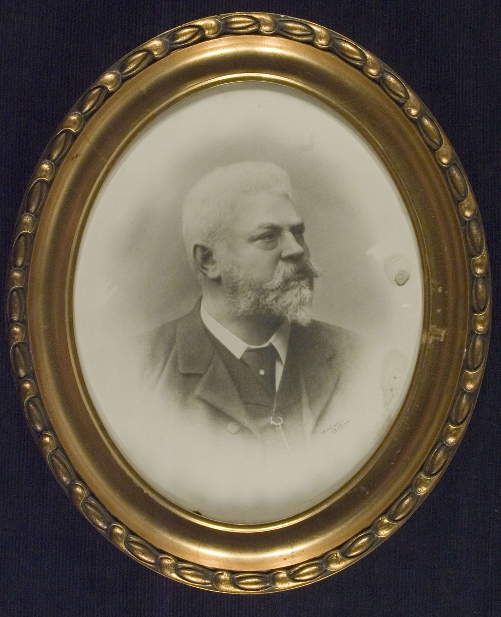 Portrett av apoteker Joachim M. Poulsen, skjegg og bart. Iført dress, hvit skjorte og slips.