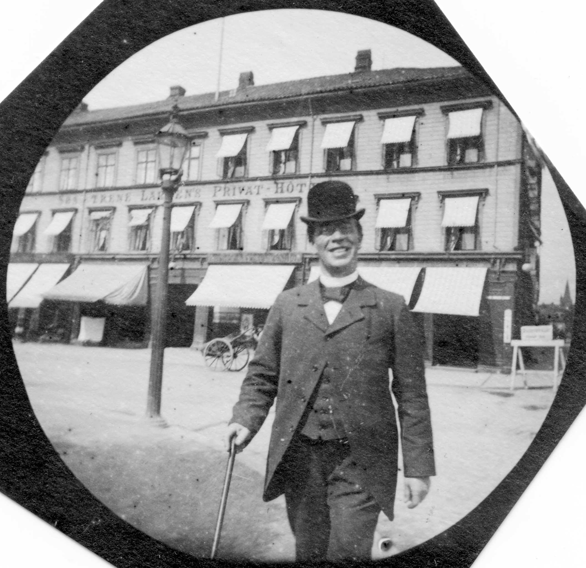 Adjunkt Kristiansen ("Kubein") med hatt, dress og stokk foran "Søstrene Larsen`s Privat-Hotel".