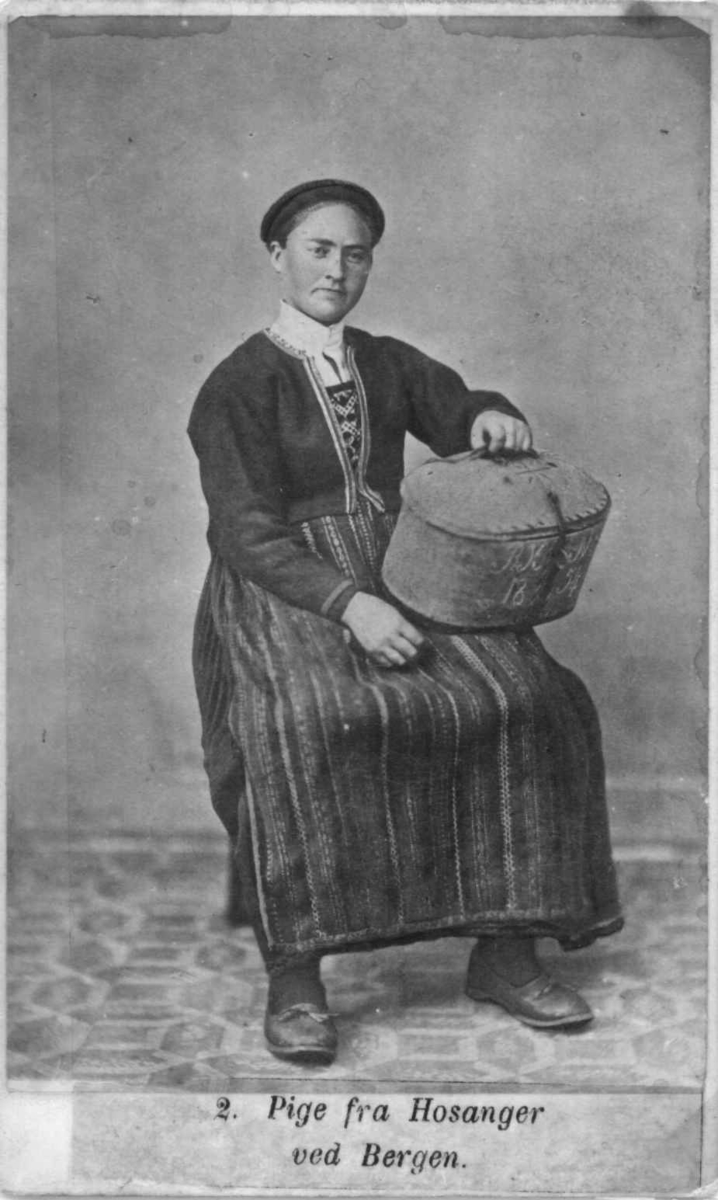 2. Jentedrakt, kvinnedrakt, Hosanger, Osterøy. Portrett av jente/kvinne sittende på stol i fotoatelier med nøytral bakgrunn og mønstret gulvteppe. Hun holder skrin/øskje (?) i fanget. Visittkortformat.
Fra Marcus Selmers (1819-1900) folkedraktserie "Norske Nationaldragter" (nr.2).