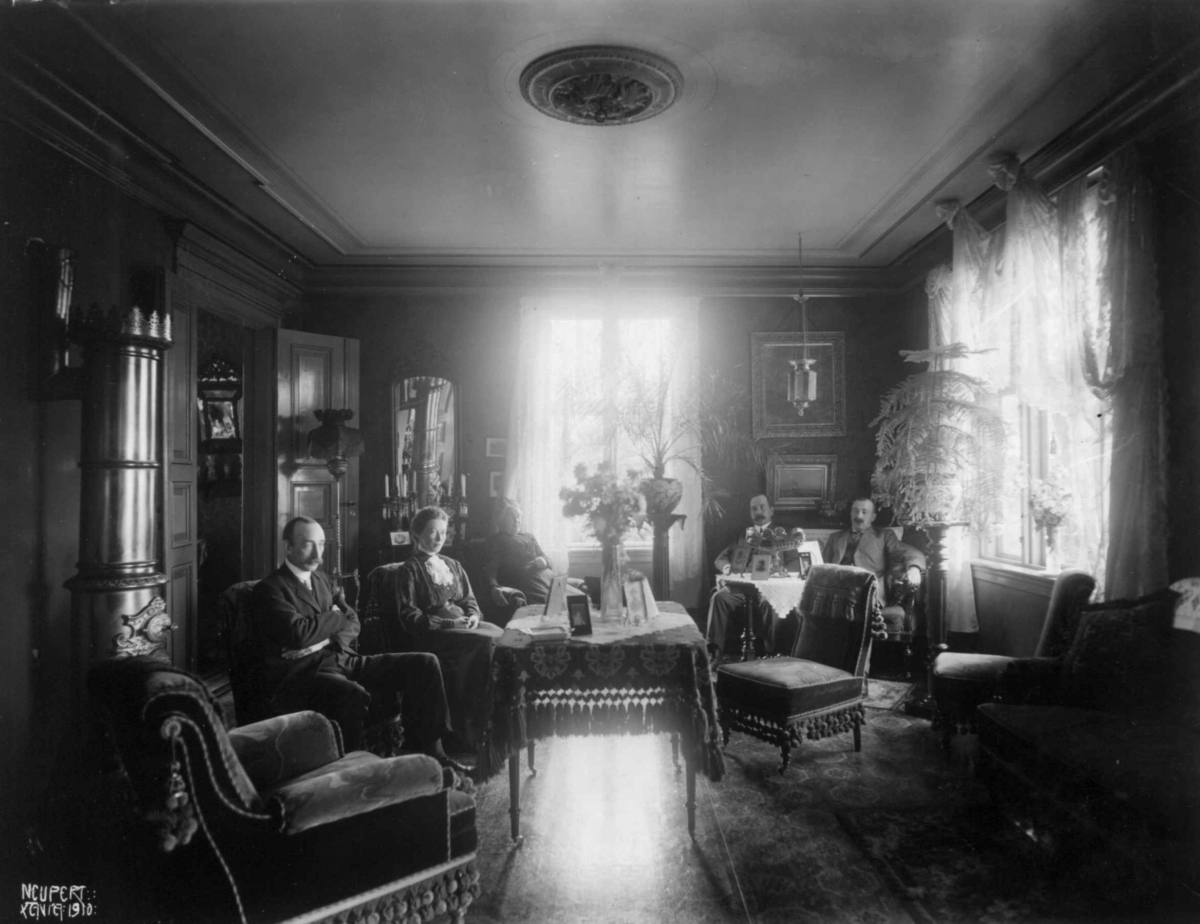 Holm. Stueinteriør. 1908-1910. To menn i en sofa. En mann og to kvinner sittende rundt et bord. Stueplanter og blomster. 