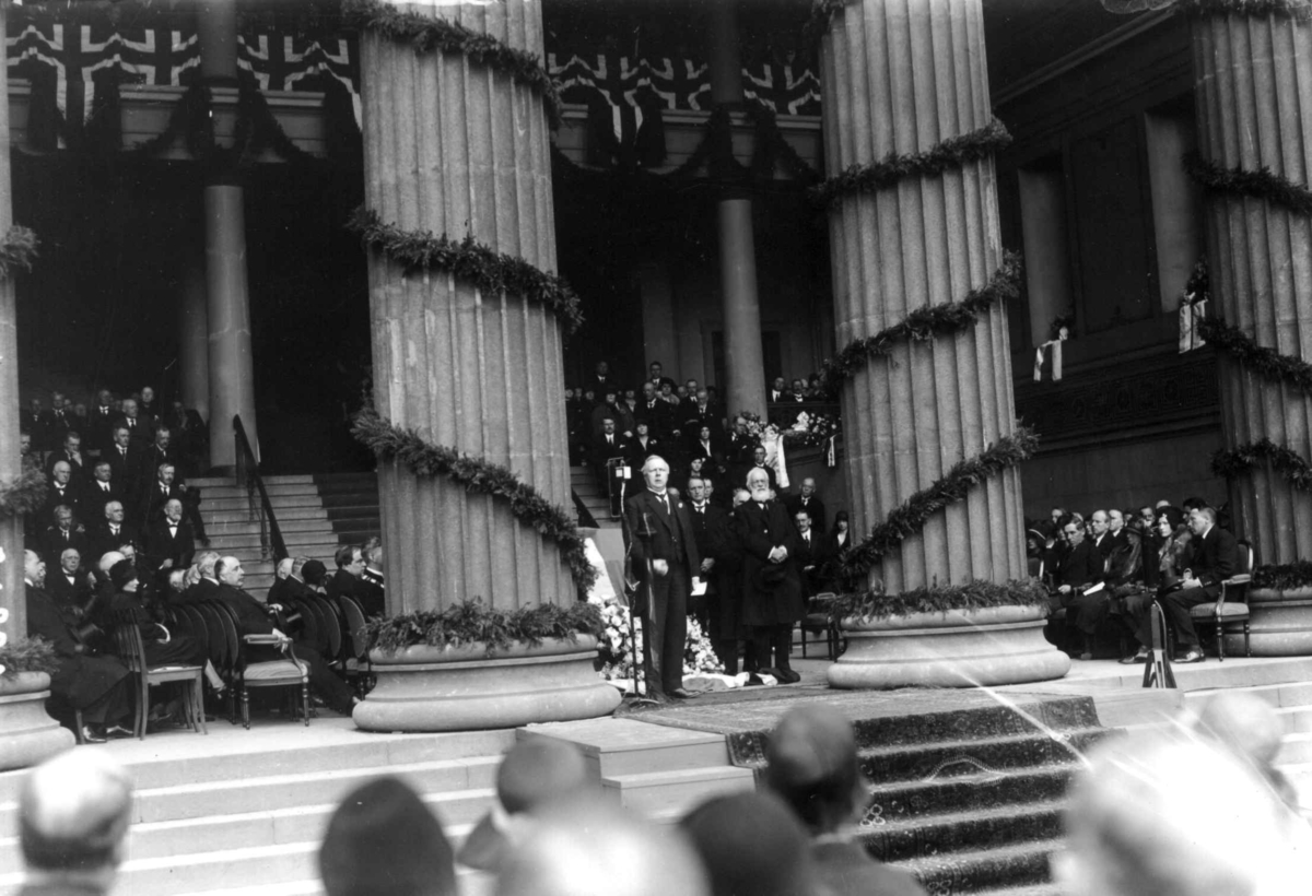 Fridtjof Nansens begravelse 17. mai 1930. Hans kiste på lit de parade i søylehallen på Universitetet, Oslo. Søylene pyntet med flagg og grønt. Æresgjester. Publikum på Universitetsplassen. 