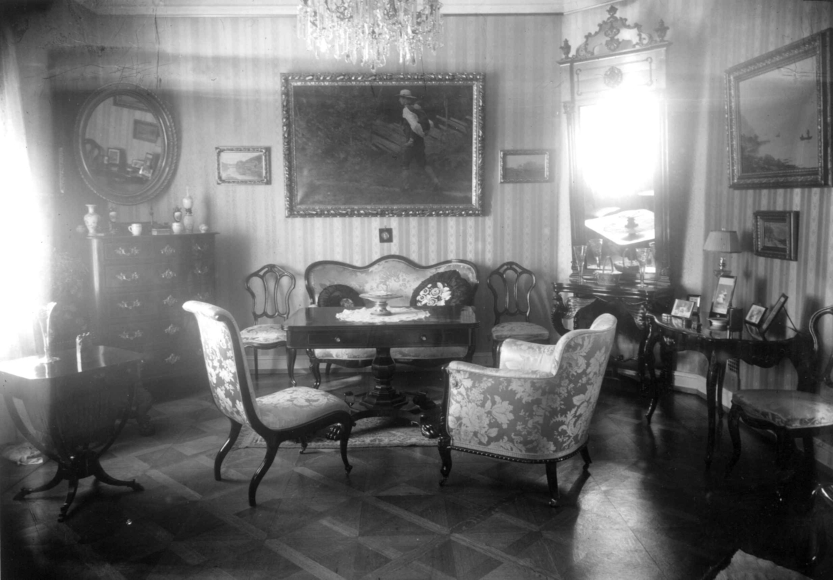 Trapp-Mayer. Stueinteriør 1925. Sofa med bord og stoler.
Speil og malerier på veggene. Lysekorne.
