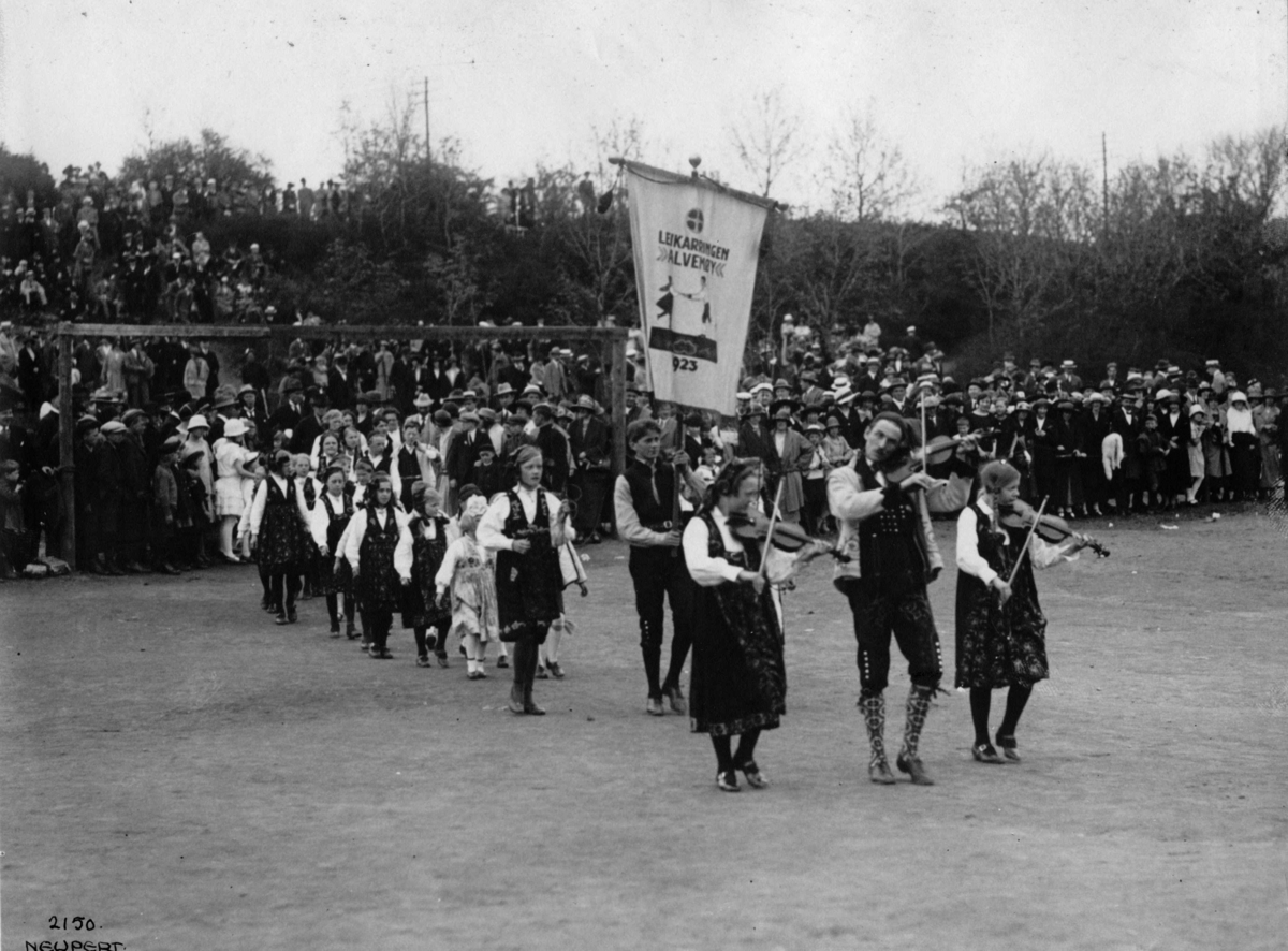 Folkedansere på vårfest på Hovedøya, Oslo 1924.
Felespillere i spissen for prosesjon av barn i bunader. Publikum.