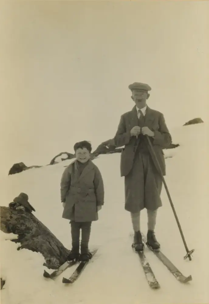 En gutt og en mann står på ski og ser inn i kameraet, de har på seg frakk og jakke i ull, og mannen har nikkers, skjorte og slips. Og bart. De står i et fjellandskap.