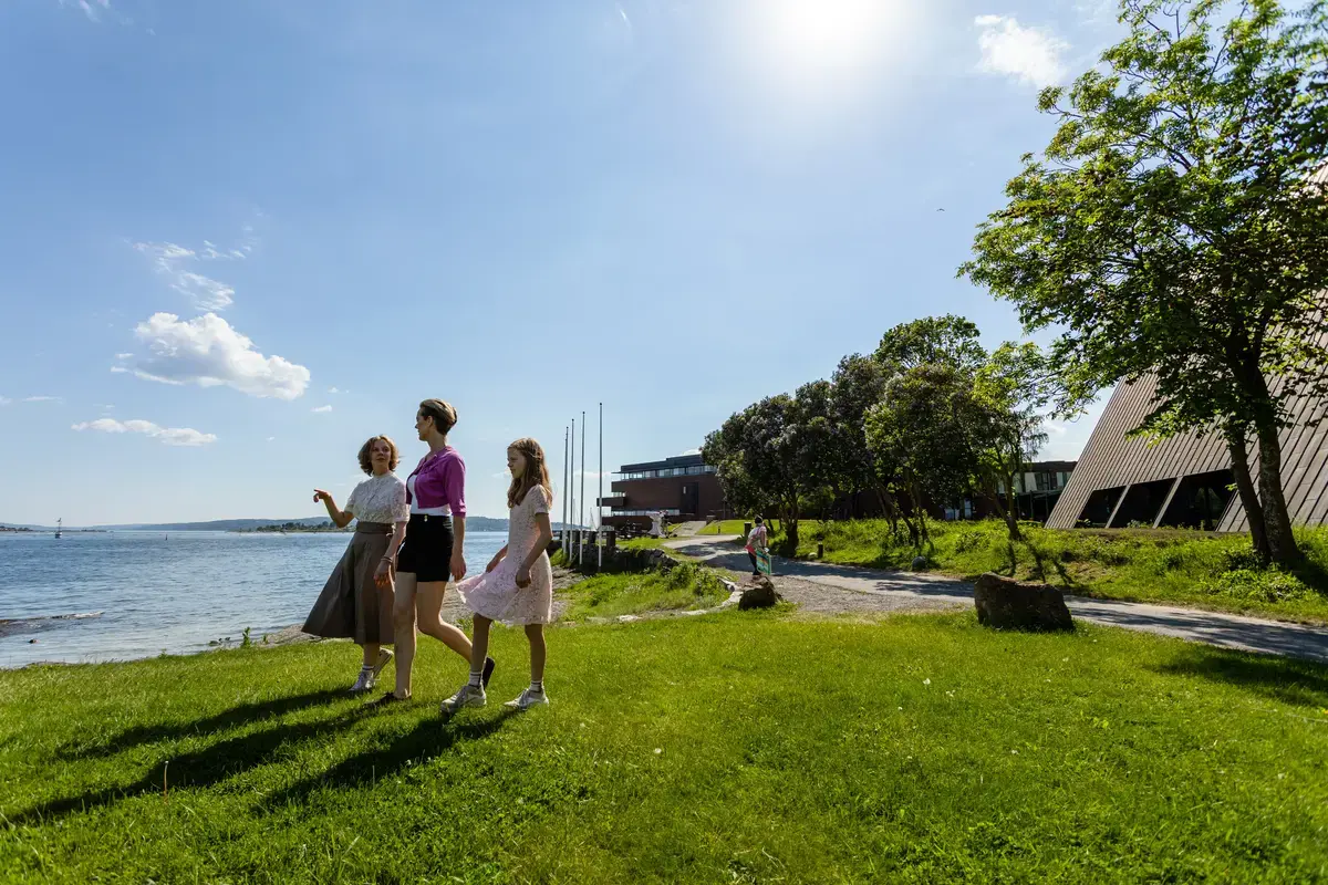 Tre personer går langs vannkanten ved Norsk Maritimt Museum ved Bygdøy i Oslo en solrik sommerdag. Norsk Maritimt Museum og Oslofjorden sees i bakgrunnen.