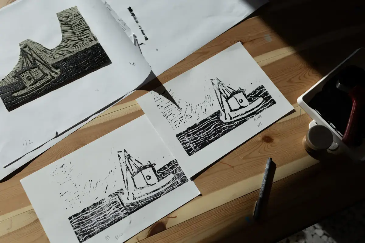Grafiske trykk av en båt i sjøen ligger på et trebord, sammen med trykkplate, rulle og trykksverte.