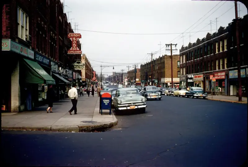 Streetscape from 'Lapskaus Boulevard' in New York, with bustling shops, passersby, and vintage cars.