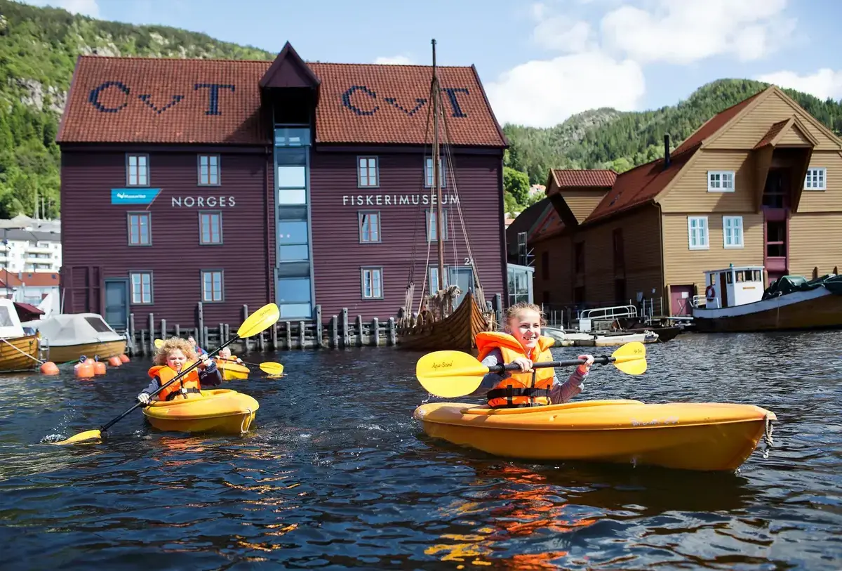 VI ser to barn som padler i hver sin gule kajakk i havnen foran Fiskerimuseet