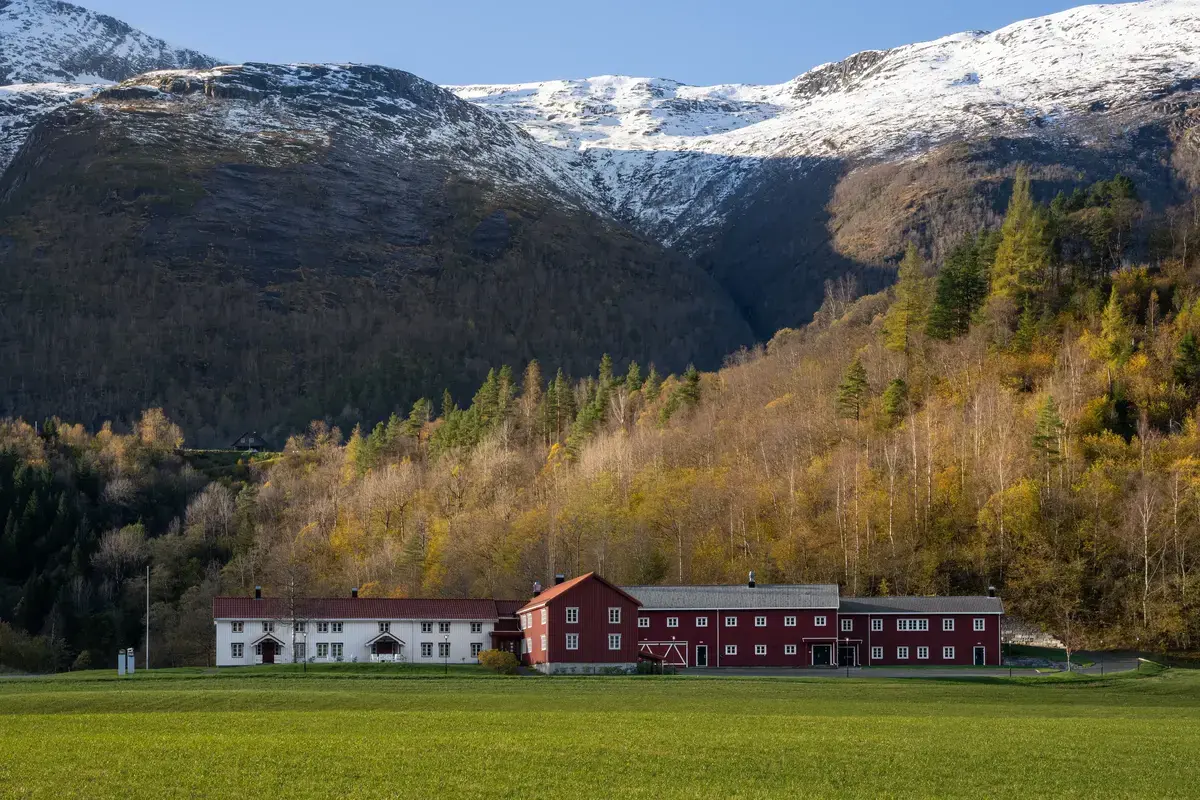 Store gårdsbygninger. Grønt jorde i forgrunn og snødekte fjell i bakgrunn.